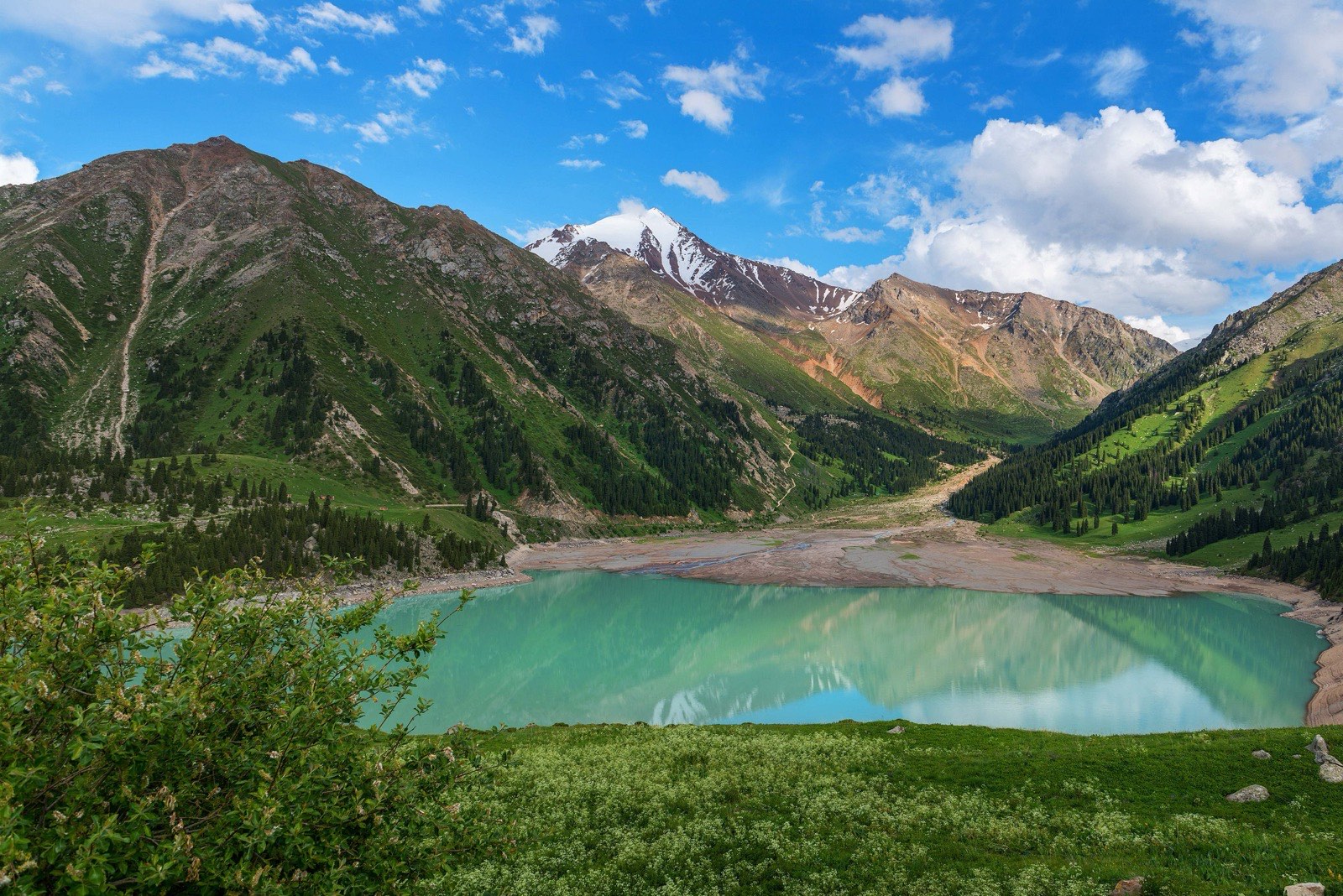 Big Almaty Lake in summer, turquoise water below green slopes and a snow-capped peak, the view that January replaces with ice and snow
